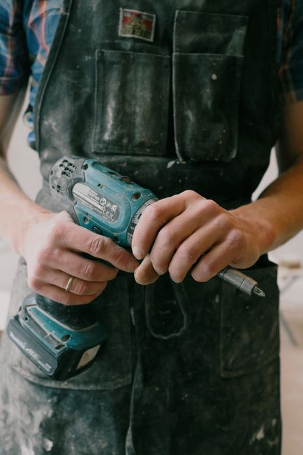 A construction worker dressed in casual white shirt and dark pants is engaged in surface cleaning at a building site, bending over and pouring a bag of cement or similar material into a white bucket p