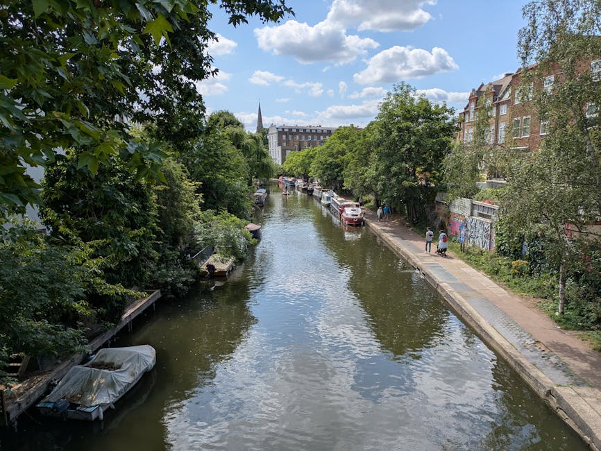 A daytime view of a narrow canal in London Fields, Hackney, bordered by a paved pathway and lush green trees on both sides. Several small boats are moored along the canal's edge, some covered with pro