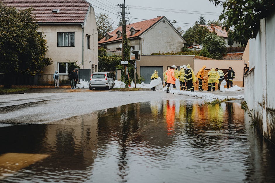 A group of emergency responders wearing yellow and orange protective suits and reflective gear working on a flooded residential street. They are handling sandbags to prevent further water intrusion, w