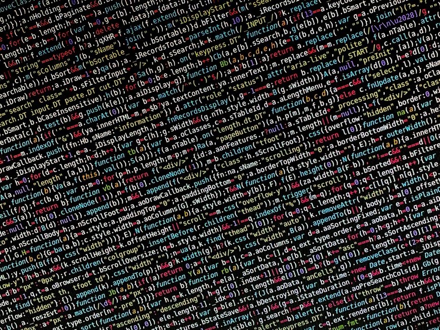 A close-up of a person's hand typing on a laptop keyboard, which displays lines of HTML and coding text on the screen. The laptop is positioned on a wooden desk with a smooth surface. The person's wri