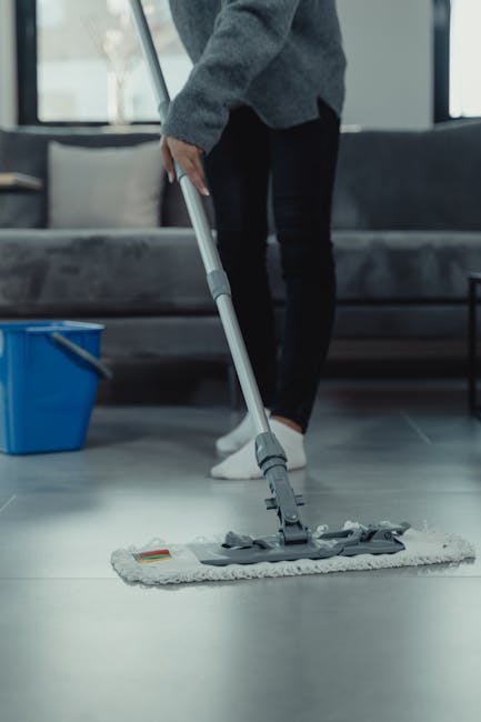 A person dressed in a grey sweater, black trousers, and white shoes is using a grey flat mop to clean a smooth, dark grey tile floor in a modern living room. The room features a grey upholstered sofa 