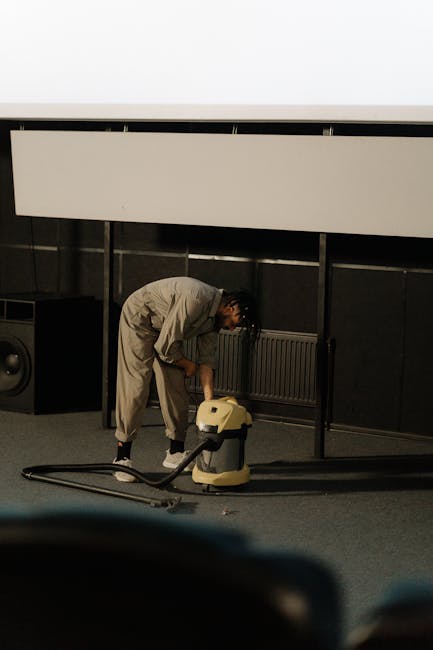 A person dressed in a grey sweater, black trousers, and white shoes is using a grey flat mop to clean a smooth, dark grey tile floor in a modern living room. The room features a grey upholstered sofa 
